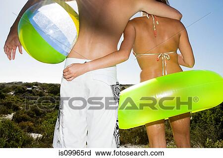 Stock Image - Young couple with inflatable ring and beach ball, rear view. Fotosearch
