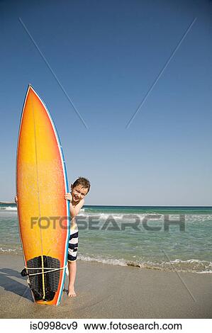 Stock Photo - Boy holding surfboard on its end in the sand. Fotosearch
