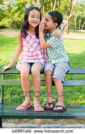 Stock Photograph - Boy whispering secrets to girl on park bench. Fotosearch