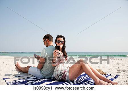 Stock Image - Couple sitting back to back on a beach while using mobile phones. Fotosearch