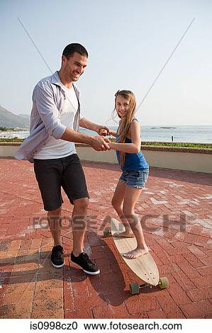 Stock Image - Father helping his daughter to ride a skateboard. Fotosearch