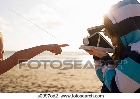 Stock Image - Girl pointing at friend with instant camera at beach. Fotosearch
