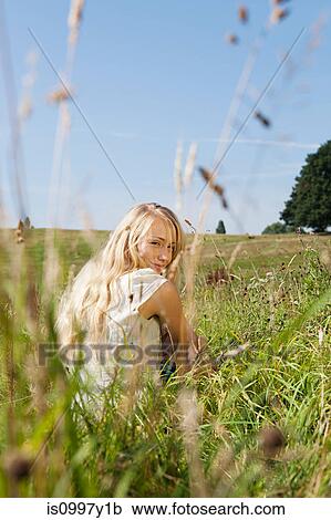 Stock Image - Young woman sitting in a field, smiling backwards at the camera. Fotosearch