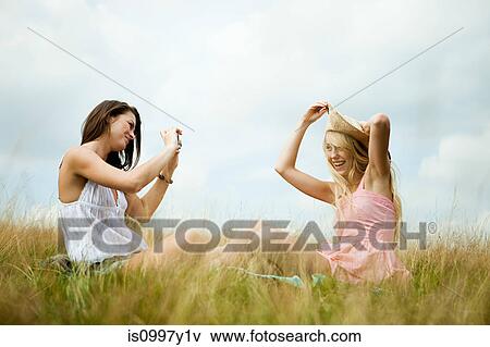 Stock Photo - Young woman taking photograph of a friend posing with a sunhat. Fotosearch