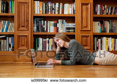 Stock Photography - Young woman using laptop by bookshelves. Fotosearch
