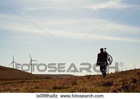 Man carrying bicycle up hill towards windfarm View Large Photo Image Stock Photo - Man carrying bicycle up hill towards windfarm. Fotosearch