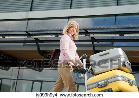 Picture - Mid adult woman pushing luggage trolley to airport. Fotosearch