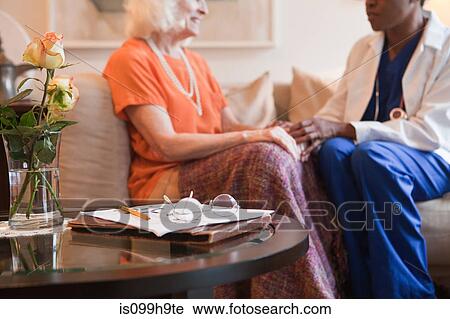 Stock Image - Senior woman holding hands with doctor in home. Fotosearch