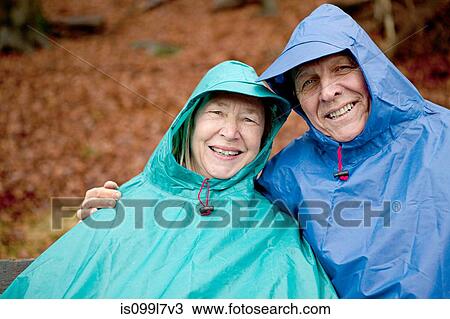 Stock Image - Portrait of senior couple sitting on park bench wearing waterproofs. Fotosearch
