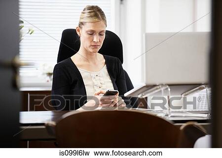 Stock Photo - Businesswoman at desk, looking at cellphone. Fotosearch