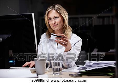 Stock Image - Businesswoman in dark office, using credit card online. Fotosearch