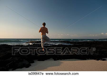 Stock Image - Man with bare chest performing yoga pose on Lava flow by the ocean on the Big Island of Hawaii. Fotosearch