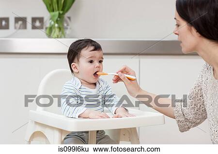 Stock Photograph - Mother feeding baby son in high chair. Fotosearch