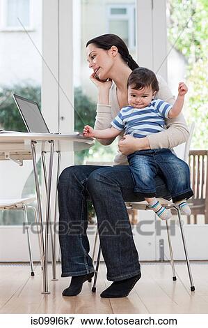 Stock Photo - Mother holding baby boy, using cellphone and laptop. Fotosearch