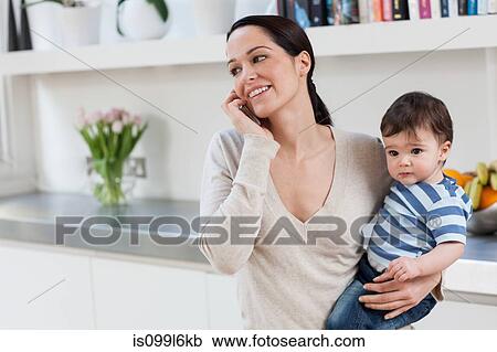 Stock Image - Mother on cellphone, holding baby son. Fotosearch