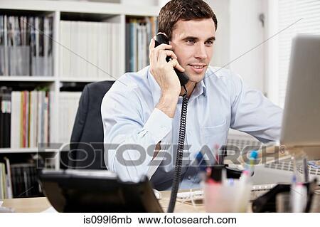 Stock Image - Young man on telephone and working on laptop. Fotosearch