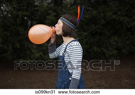 Stock Image - Young woman in headdress, blowing up a balloon. Fotosearch