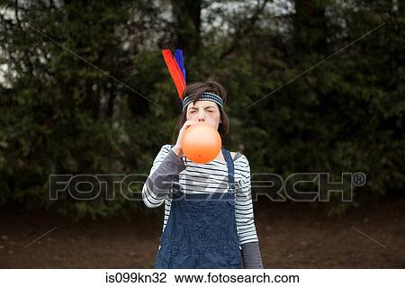 Stock Image - Young woman in headdress, blowing up a balloon. Fotosearch