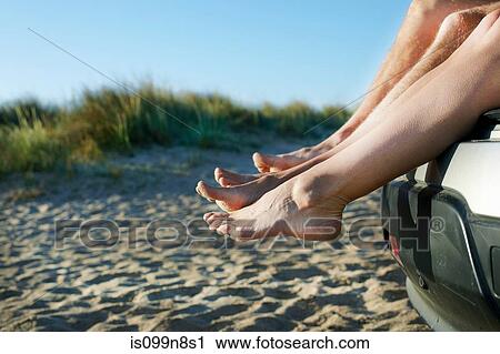 Stock Image - Mid adult couple sitting barefoot on car on beach. Fotosearch
