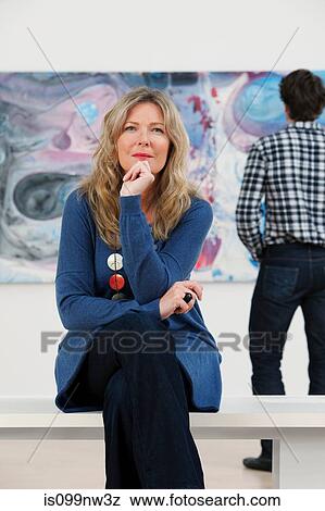 Stock Photo - Mid adult woman sitting in front of oil painting in gallery. Fotosearch