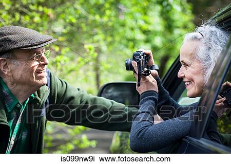 Stock Image - Senior woman photographing man from car. Fotosearch