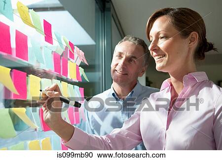 Stock Image - Two businesspeople writing on adhesive notes on window. Fotosearch