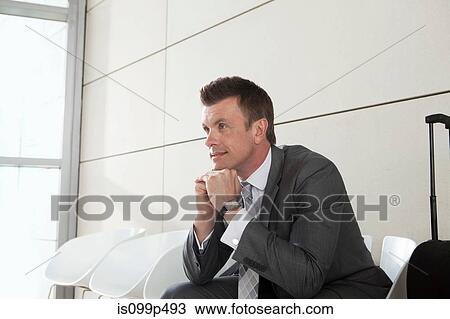 Stock Image - Businessman waiting with luggage, hand on chin. Fotosearch