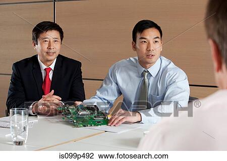 Stock Image - Businessmen discussing circuit board in meeting. Fotosearch