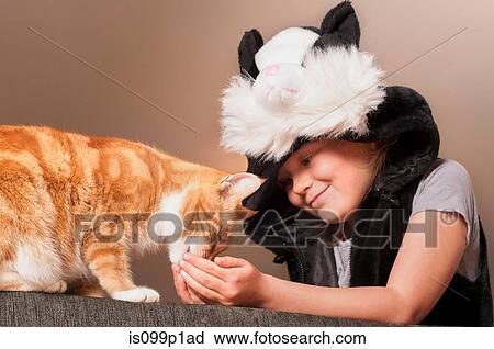 Stock Image - Girl in cat costume feeding pet cat. Fotosearch