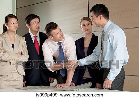 Stock Photo - Multi racial businesspeople shaking hands over blueprint. Fotosearch
