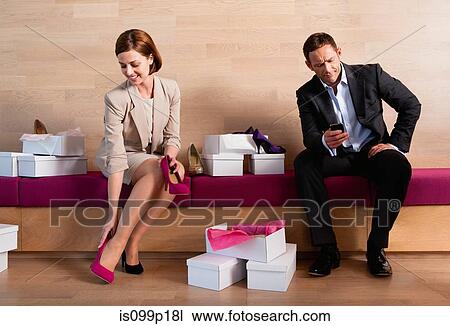 Stock Image - Woman trying on shoes in shoe shop, bored man texting. Fotosearch