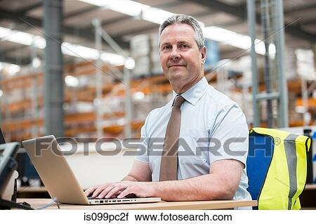 Stock Image - Man using laptop in warehouse, portrait. Fotosearch
