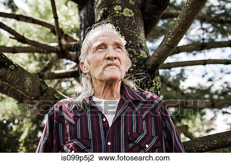 Stock Image - Senior man by tree trunk, looking away. Fotosearch