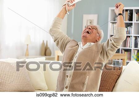 Stock Image - Senior man holding remote control cheering. Fotosearch