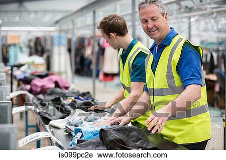 Stock Image - Two men sorting clothes on conveyor belt in warehouse. Fotosearch