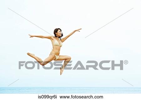 Stock Image - Young woman in bikini jumping on beach. Fotosearch
