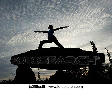 Stock Image - Silhouette of woman in warrior yoga pose on a rock. Fotosearch