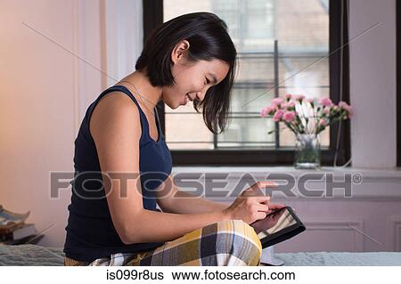 Young woman sitting on bed, using digital tablet View Large Photo Image Stock Photograph - Young woman sitting on bed, using digital tablet. Fotosearch