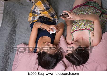 Young women lying on bed, looking at cellphones, overhead view View Large Photo Image Stock Photography - Young women lying on bed, looking at cellphones, overhead view. Fotosearch