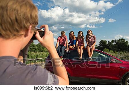 Stock Image - Friends sitting on car roof, young man taking photograph. Fotosearch