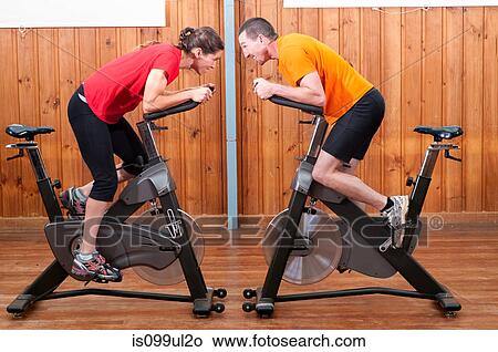 Picture - Man and woman competing against each other on exercise bikes. Fotosearch