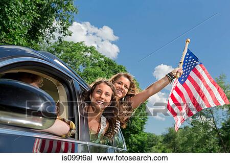 Stock Image - Two friends leaning out of car window holding american flag. Fotosearch