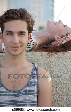 Stock Photograph - Young woman lying on wall with young man in front, portrait. Fotosearch