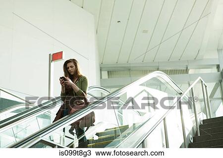 Stock Image - Young woman on escalator using smartphone. Fotosearch