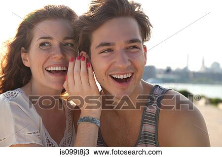 Stock Image - Young woman whispering to boyfriend. Fotosearch