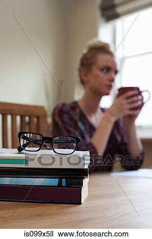 Stock Image - Books and pair of glasses in foreground, young woman with coffee cup in background. Fotosearch