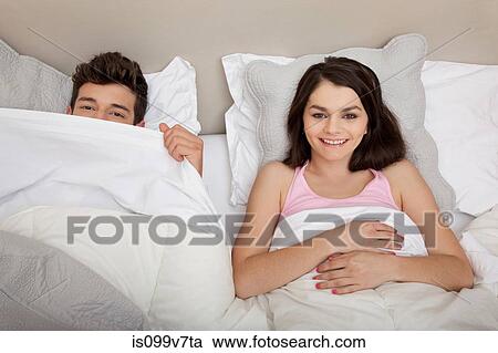 Stock Image - Couple in bed, man peeking behind duvet. Fotosearch