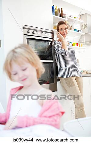 Mother on cell phone in kitchen with daughter in foreground View Large Photo Image Picture - Mother on cell phone in kitchen with daughter in foreground. Fotosearch