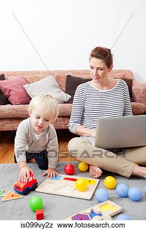 Stock Photo - Mother using laptop, son playing. Fotosearch