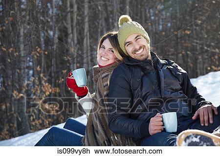 Stock Image - Portrait of couple sitting in snow with hot drinks. Fotosearch